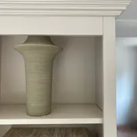 Detail of the top corner, showing the stepped moulded cornice and the frame of the off-white shelving with a ribbed green vase standing on the shelf below