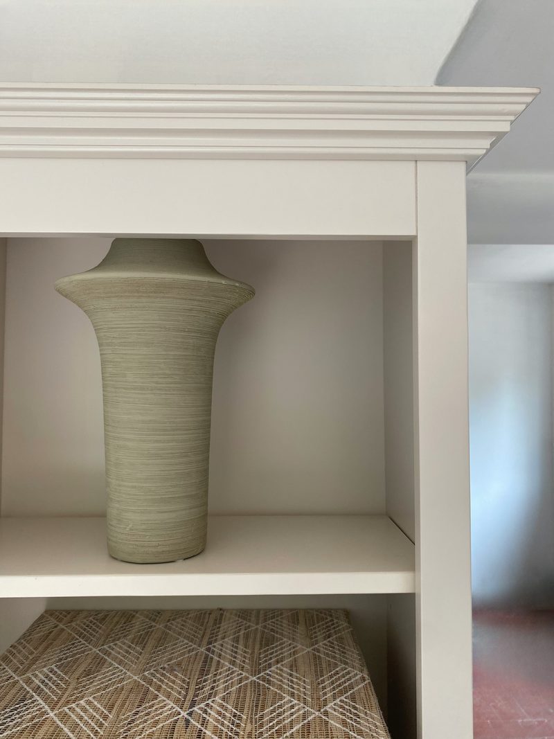 Detail of the top corner, showing the stepped moulded cornice and the frame of the off-white shelving with a ribbed green vase standing on the shelf below