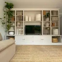 Angled view of the media wall from the left, with the arm of a beige sofa in the foreground, the television centred on the cupboard top and a plant trailing down the end shelves