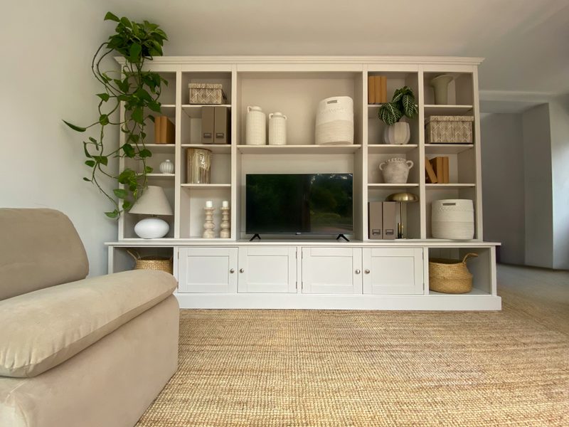 Angled view of the media wall from the left, with the arm of a beige sofa in the foreground, the television centred on the cupboard top and a plant trailing down the end shelves