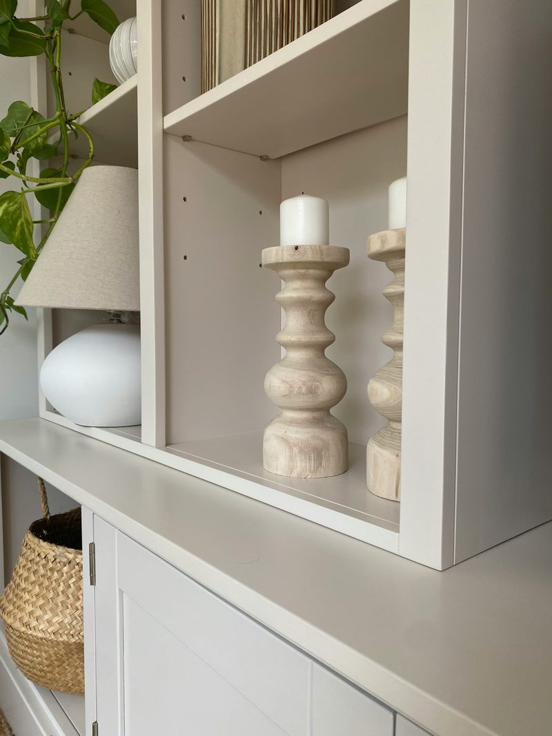 Close view of two turned pale wood candlesticks with white pillar candles on an open shelf, beside a white ceramic lamp, above the cupboard top and a seagrass basket