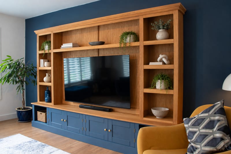Angled view of the Woburn media unit showing the oak top section with open shelves styled with plants, books and ceramics above navy painted cupboards with brass knobs