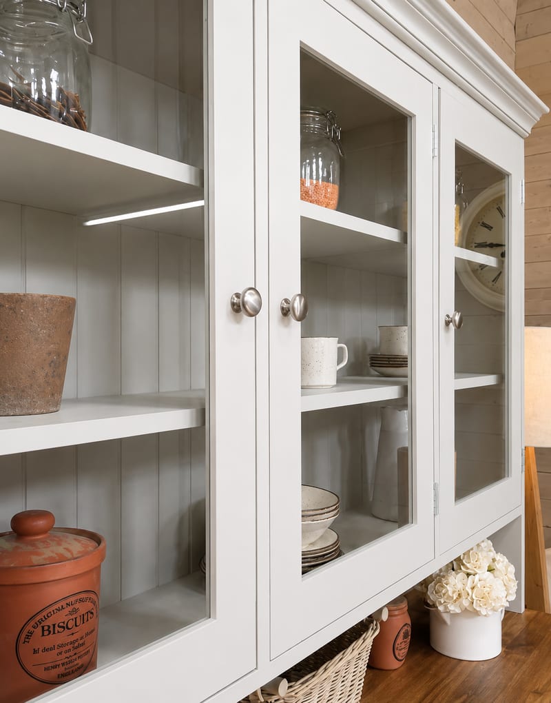 Close view of the glazed upper doors of a cream painted Welsh dresser, showing shelves of mugs, bowls and storage jars behind the glass with a stoneware biscuit barrel below