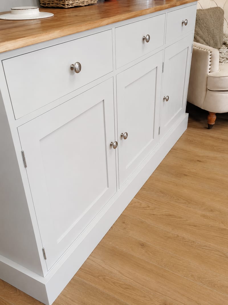 Angled view of the painted base unit from the side, showing three drawers over three panelled doors, brushed nickel knobs and the oak top, with a cream armchair on wood effect flooring