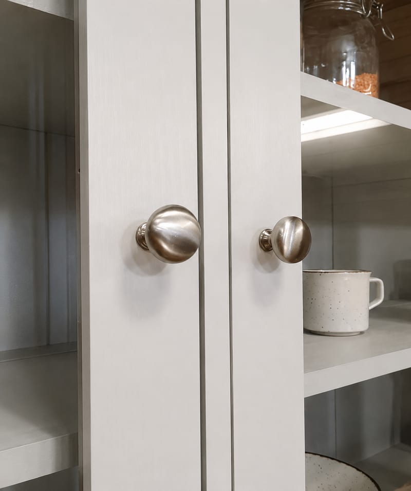 Close-up of two brushed nickel round knobs on the meeting stiles of the glazed dresser doors, with a speckled cream mug and grey painted shelves visible through the glass behind