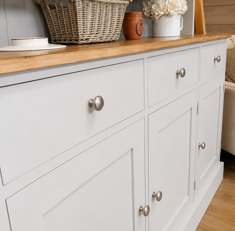 Low angled view along the oak worktop and painted drawer fronts, showing the grain of the timber top, brushed nickel knobs and a wicker basket, jug of white flowers and a stoneware jar above