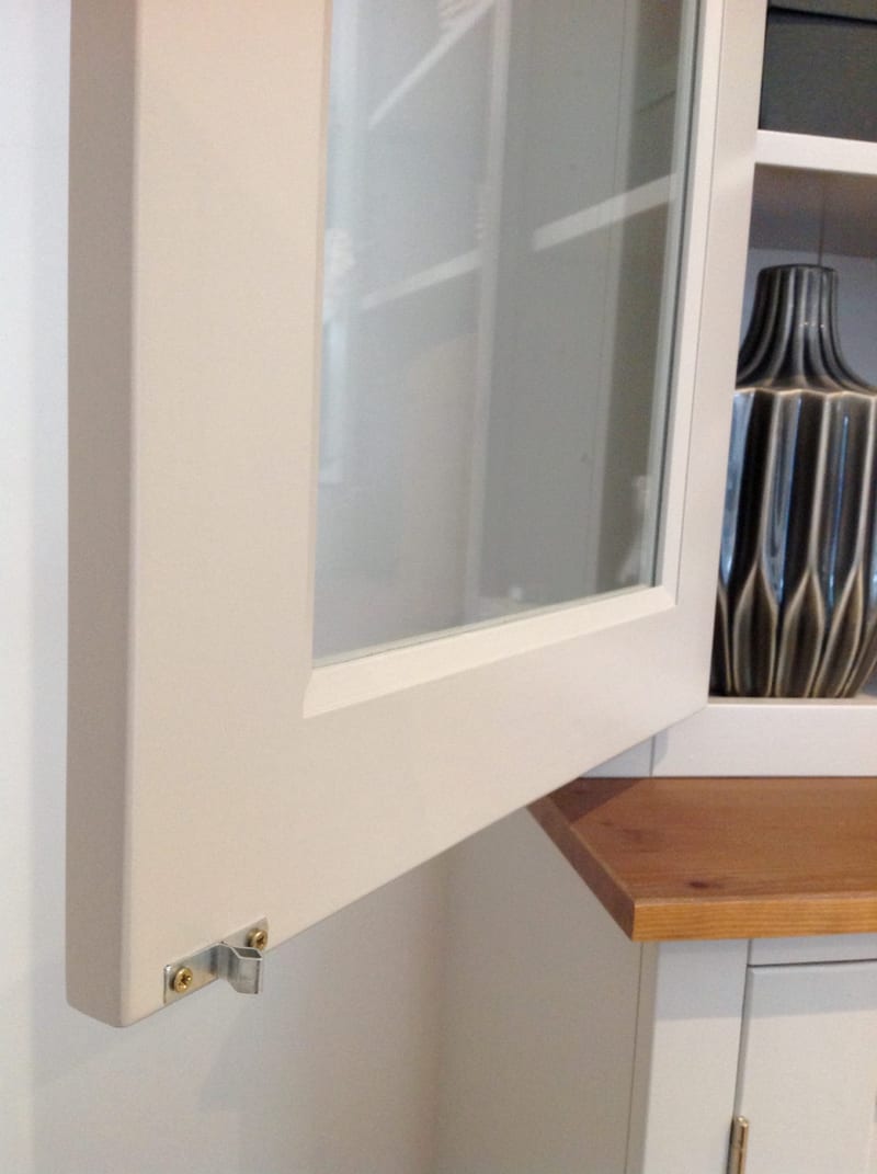 Close view of an open glazed door on the painted dresser top, showing the door edge, a steel catch plate with brass screws and a ribbed grey vase on the shelf above the pine worktop