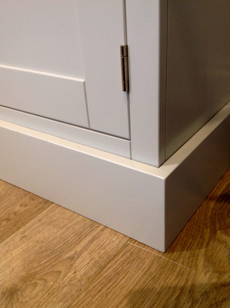 Corner detail at the base of the grey painted dresser, showing the bottom of a panelled door, its steel hinge and the stepped plinth meeting a wood effect floor
