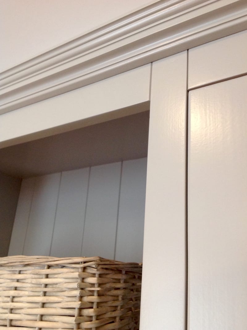 Close view of the moulded cornice and door stile on the painted dresser, with the grey tongue-and-groove back panel of the open shelf recess and a wicker basket below