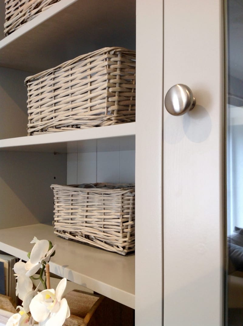 Close-up of a brushed nickel knob on the edge of a glazed door, with grey wicker storage baskets sitting on the painted shelves inside and a white orchid below