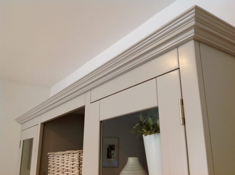 Upward angled view of the moulded cornice on the painted dresser, showing the mitred return at the corner, a steel door hinge, the room reflected in the glazed door and a wicker basket below