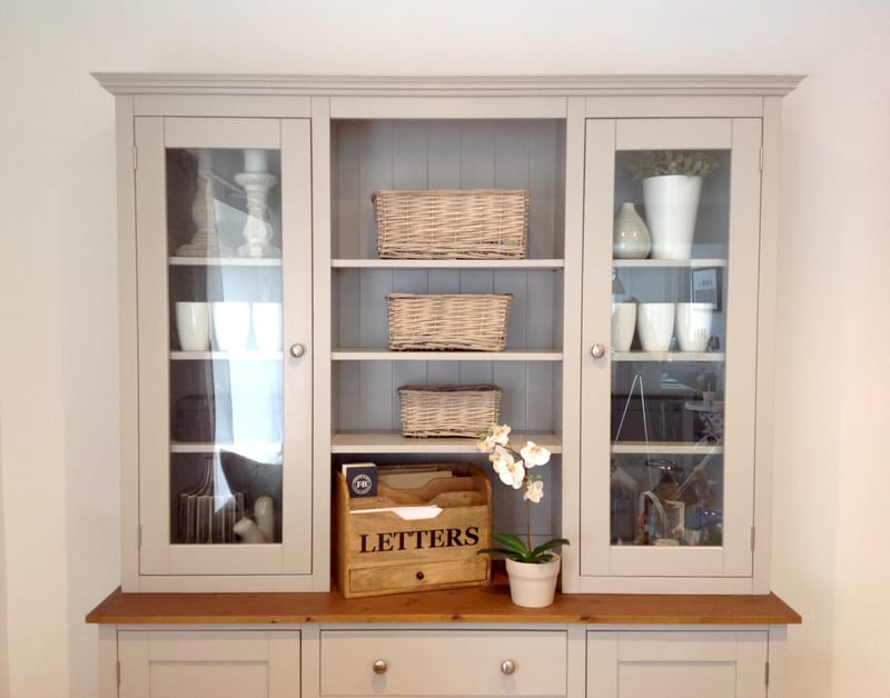 Straight-on view of the glazed top section, showing two glazed doors flanking three open shelves with wicker baskets, a wooden letters box, an orchid and white ceramics inside