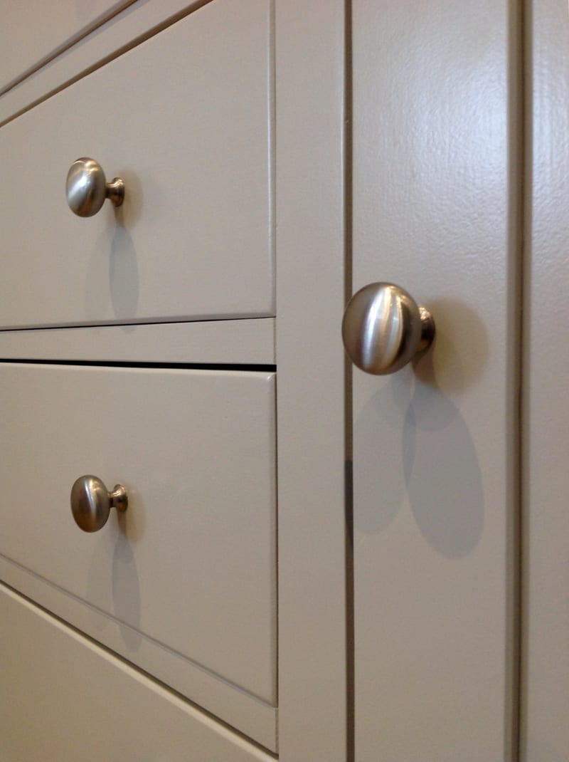 Close-up of three brushed nickel knobs on the painted drawer and door fronts, showing the rounded knob profile and the shadow gaps between each drawer front