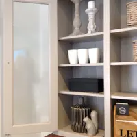 Glazed door open on the left of the dresser top, showing four painted shelves holding stone candlesticks, white vases, a dark box and a ceramic squirrel above the pine worktop
