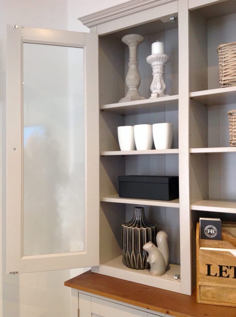 Glazed door open on the left of the dresser top, showing four painted shelves holding stone candlesticks, white vases, a dark box and a ceramic squirrel above the pine worktop