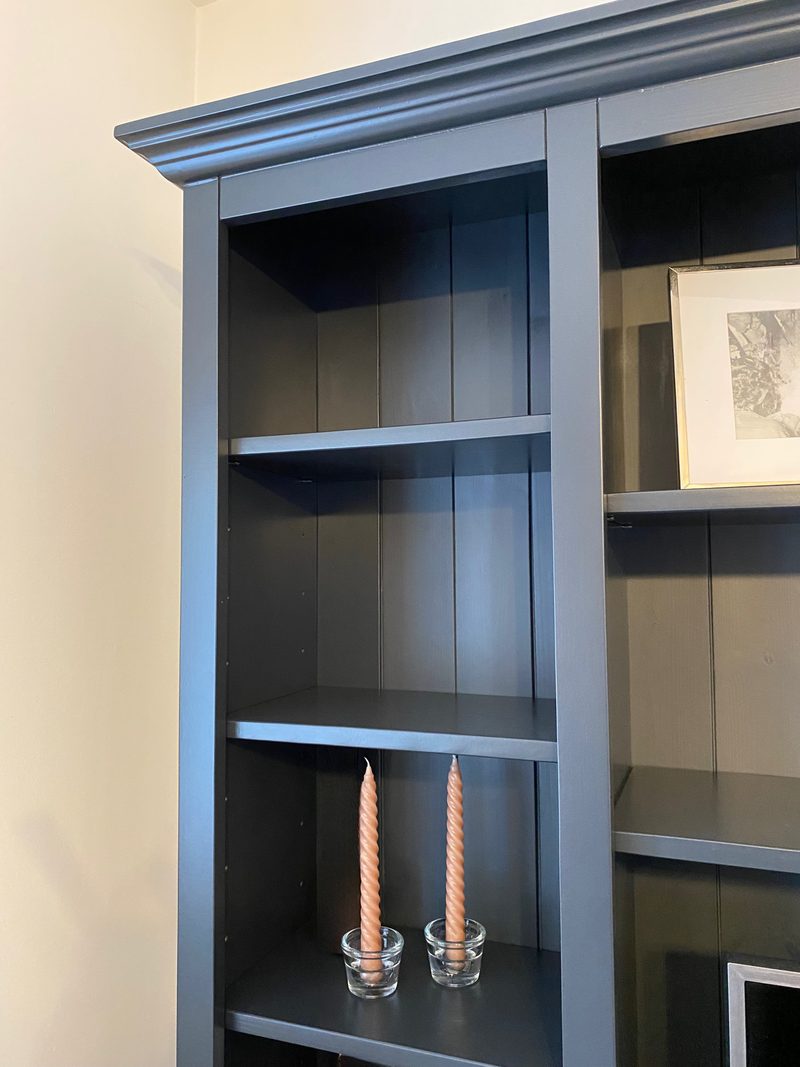 Close view of the left bookcase column of the dark painted unit, showing the moulded cornice, adjustable shelves, tongue-and-groove back panel and two twisted peach candles in glass holders