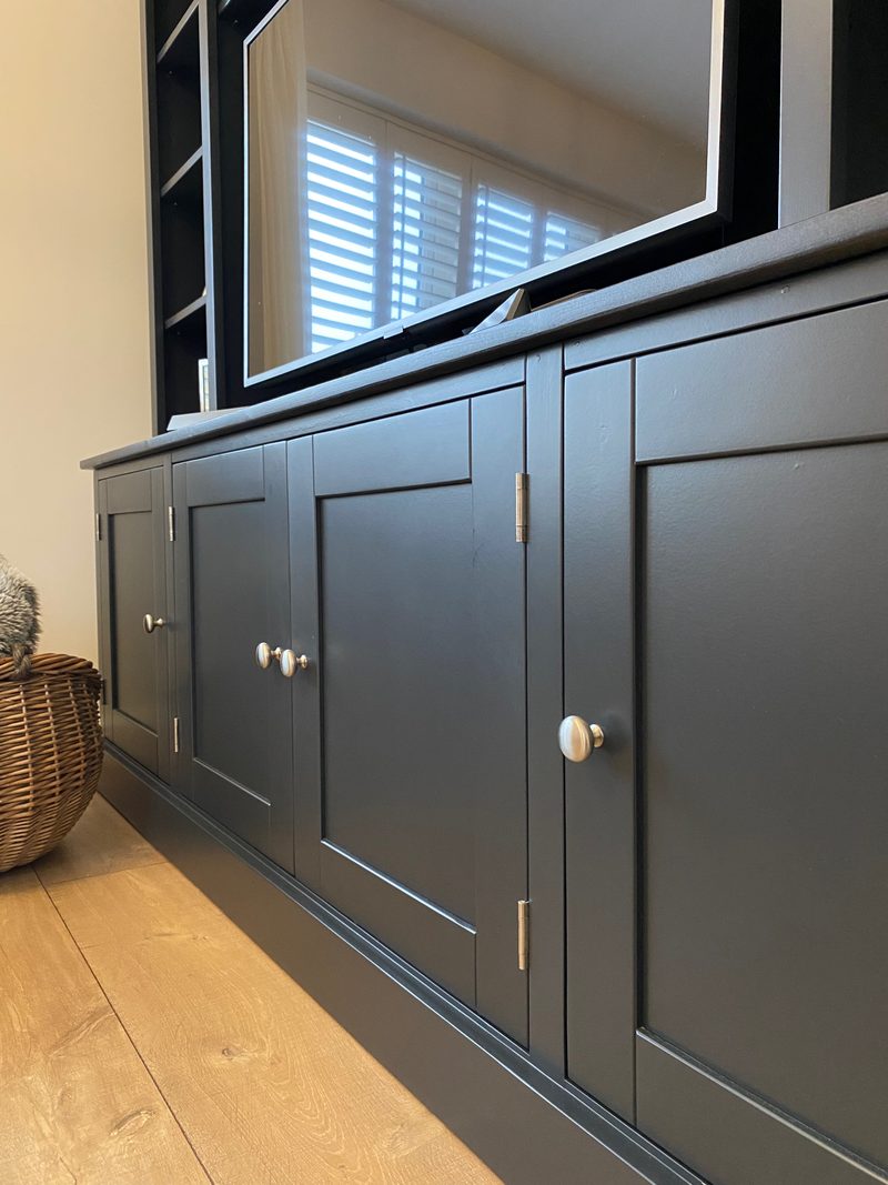 Low angled view along the base cupboard doors, showing four panelled doors with brushed nickel knobs and hinges above a stepped plinth, with the television screen reflecting window shutters
