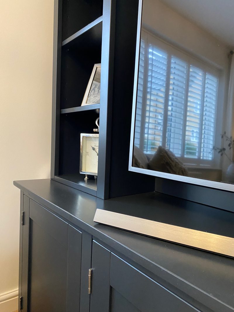 Close view of the television standing on the dark painted top, showing the shelf upright beside it with a mantel clock and framed print, and a cupboard door hinge below