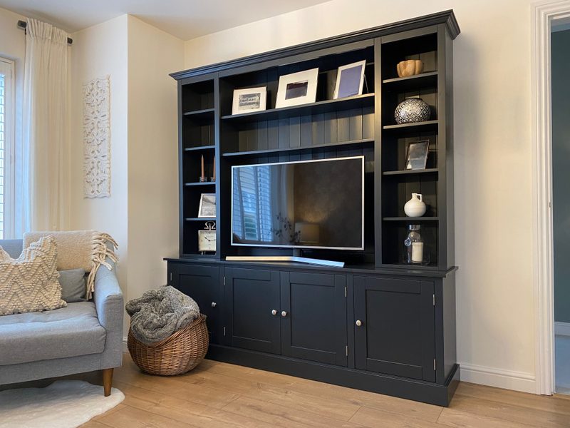 Angled room view of the full media unit against a cream wall, with a grey sofa, wicker basket and carved wall panel alongside it and a curtained window on the left