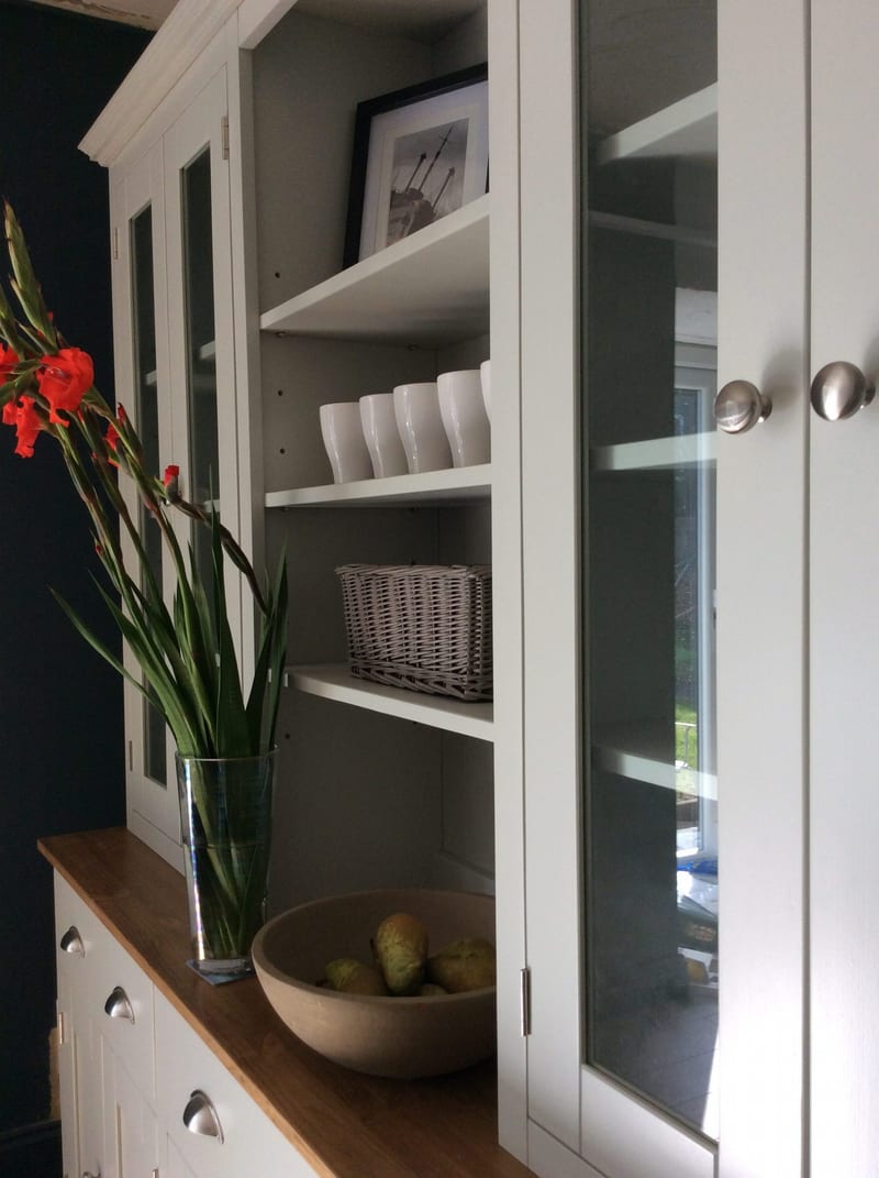 Close view from the side of the open central shelves, showing white mugs, a grey wicker basket, a framed photograph and a stone bowl of pears on the oiled timber top beside red gladioli