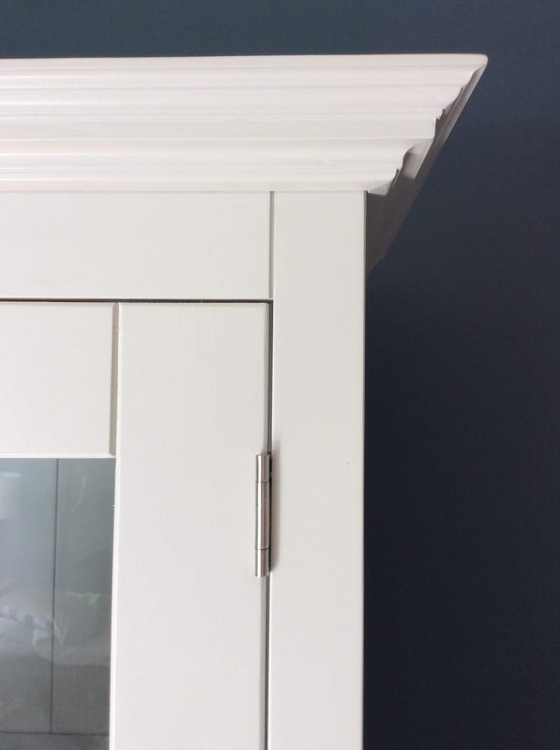 Detail of the top corner of the painted dresser, showing the layered moulded cornice, the brushed nickel butt hinge on a glazed door and the deep navy wall behind