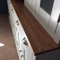 Close view along the oiled timber top from one end, showing the warm grain and squared edge above the painted drawer fronts with brushed nickel cup handles and the plank wood floor
