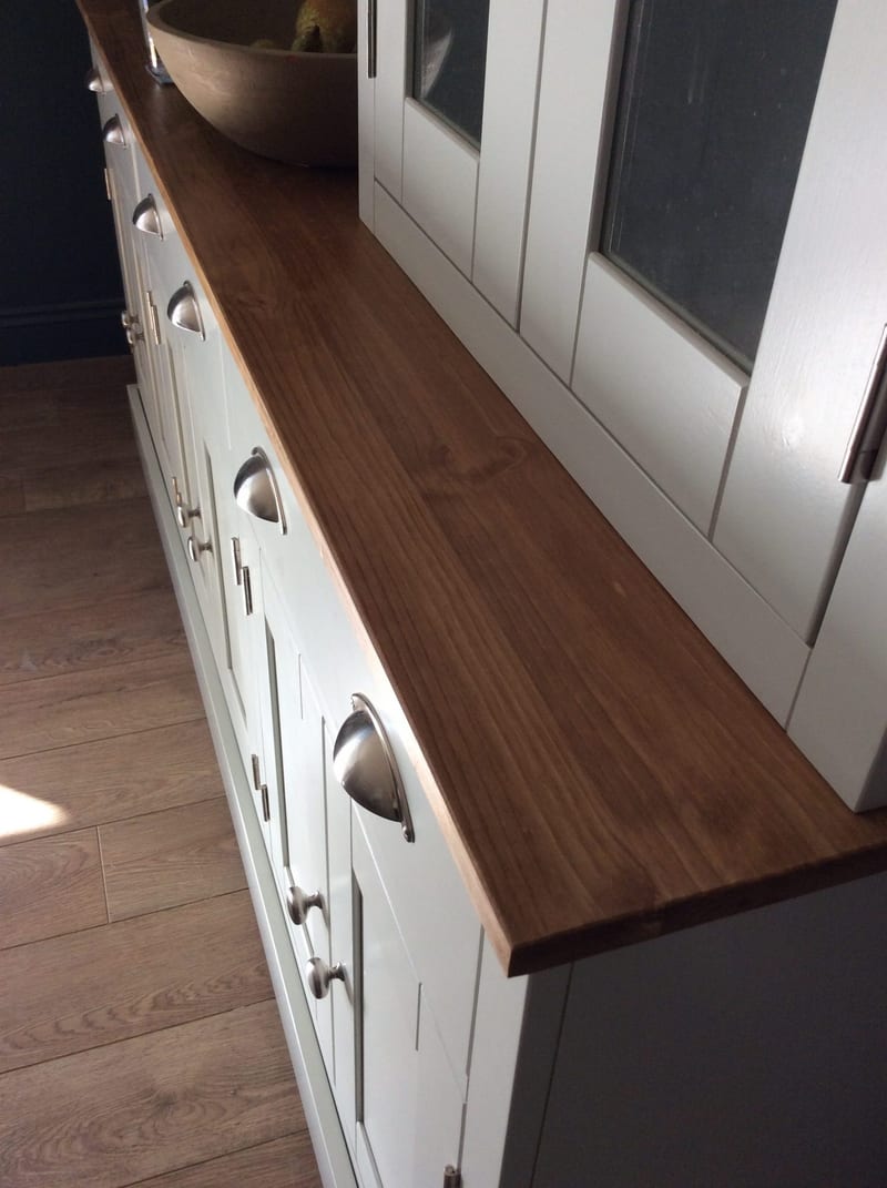 Close view along the oiled timber top from one end, showing the warm grain and squared edge above the painted drawer fronts with brushed nickel cup handles and the plank wood floor