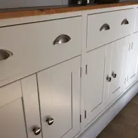 Close angled view of the base from the left, showing three drawers with brushed nickel cup handles, panelled doors with round knobs, the timber top edge and the deep painted plinth