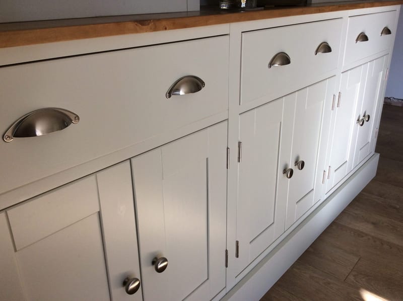 Close angled view of the base from the left, showing three drawers with brushed nickel cup handles, panelled doors with round knobs, the timber top edge and the deep painted plinth