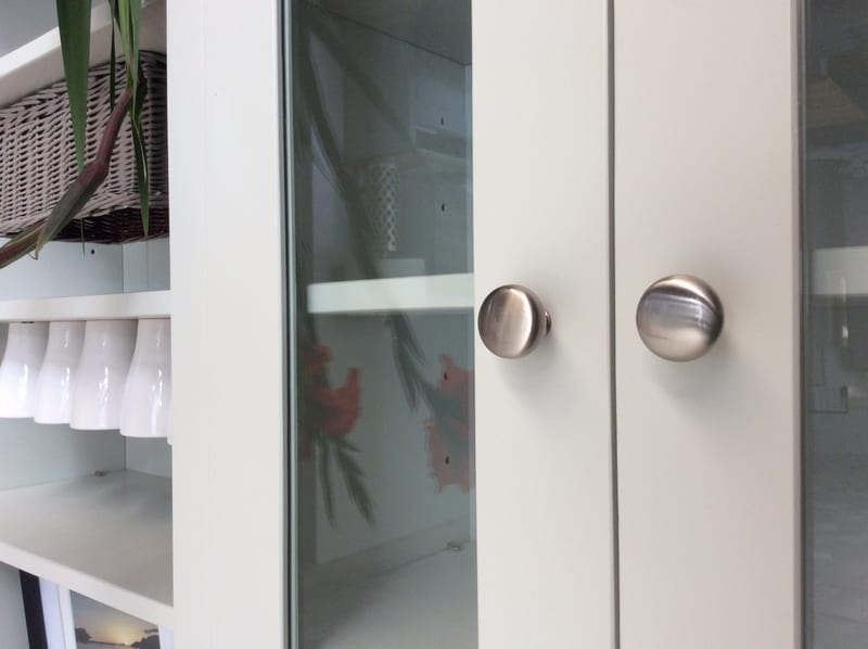 Close-up of a pair of glazed doors with round brushed nickel knobs, showing white mugs, a wicker basket and green leaves on the open painted shelves alongside and red flowers reflected in the glass