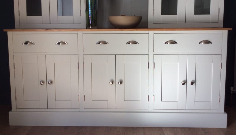 Straight-on view of the base cabinet, showing three drawers with brushed nickel cup handles, six panelled doors with round knobs, a stone bowl on the oiled timber top and the plinth on a wood floor