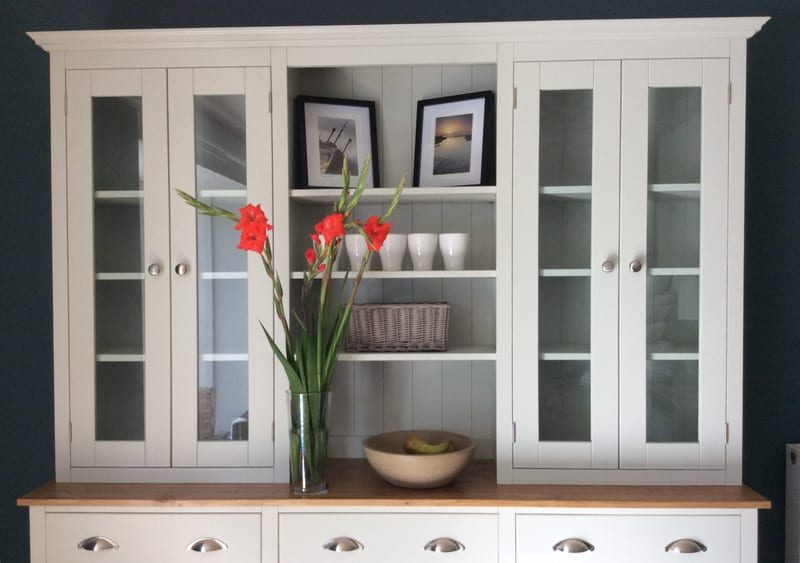 Front view of the glazed upper section, showing four glazed doors, open central shelves with two framed coastal photographs, white mugs and a wicker basket against a navy wall