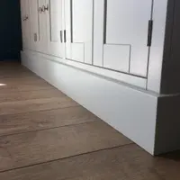 Low corner view of the painted plinth and lower door panels, showing round brushed nickel knobs, a slim hinge and the plank wood flooring running away from the piece