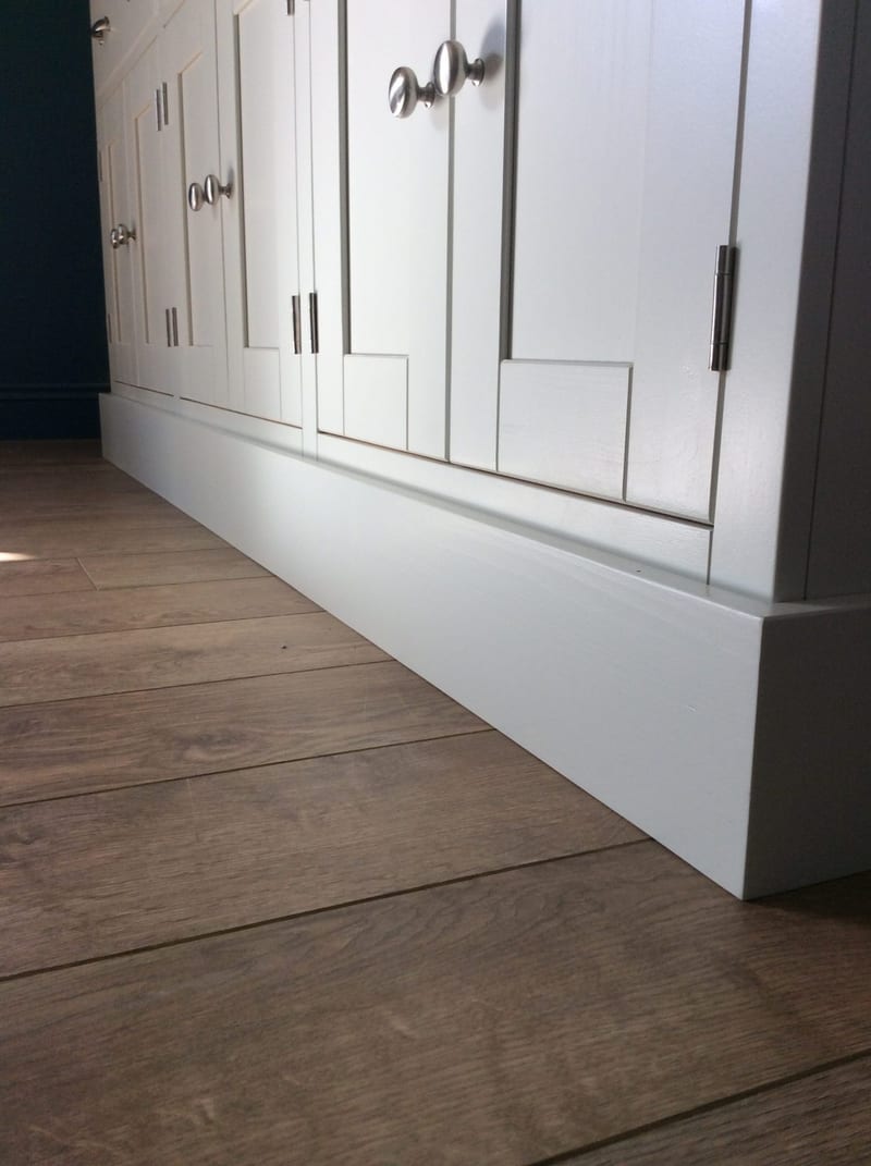 Low corner view of the painted plinth and lower door panels, showing round brushed nickel knobs, a slim hinge and the plank wood flooring running away from the piece