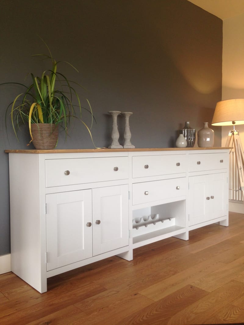 Angled view from the left of the white painted sideboard, showing the oiled timber top, drawers with round brushed nickel knobs, panelled doors and the open centre wine rack on an oak floor