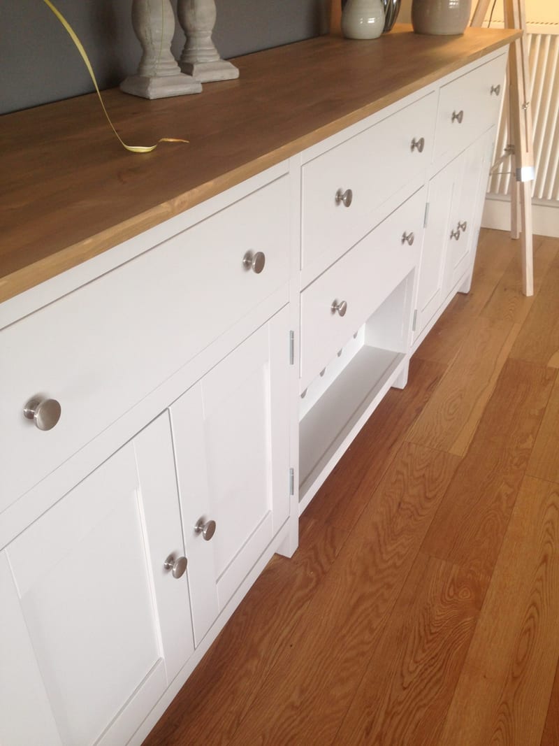 Close view along the oiled timber top from one end, showing the grain and rounded edge beside stone candlesticks, with round brushed nickel knobs on the drawers and the wine rack shelf below