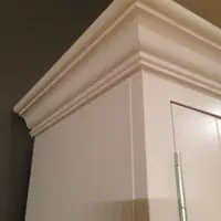 Detail of the top corner of the dresser, showing the deep layered cornice moulding, the painted side panel and a slim metal hinge on the door beside a dark grey wall