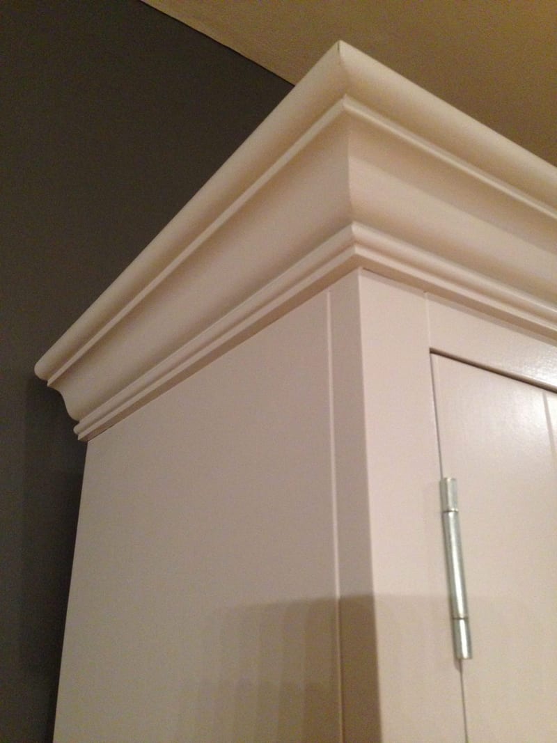 Detail of the top corner of the dresser, showing the deep layered cornice moulding, the painted side panel and a slim metal hinge on the door beside a dark grey wall