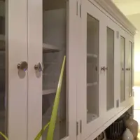 Angled view along the glazed upper section, showing six glazed doors with round brushed nickel knobs, painted interior shelves holding a wicker basket and green leaves in the foreground