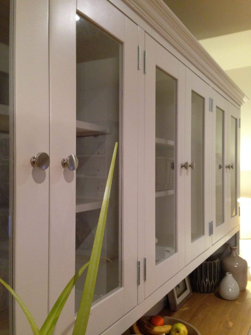 Angled view along the glazed upper section, showing six glazed doors with round brushed nickel knobs, painted interior shelves holding a wicker basket and green leaves in the foreground