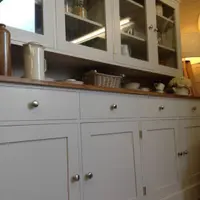 Angled view along the pale grey painted dresser showing four drawers and four cupboard doors with brushed nickel knobs, a waxed pine worktop and the glazed cabinet above