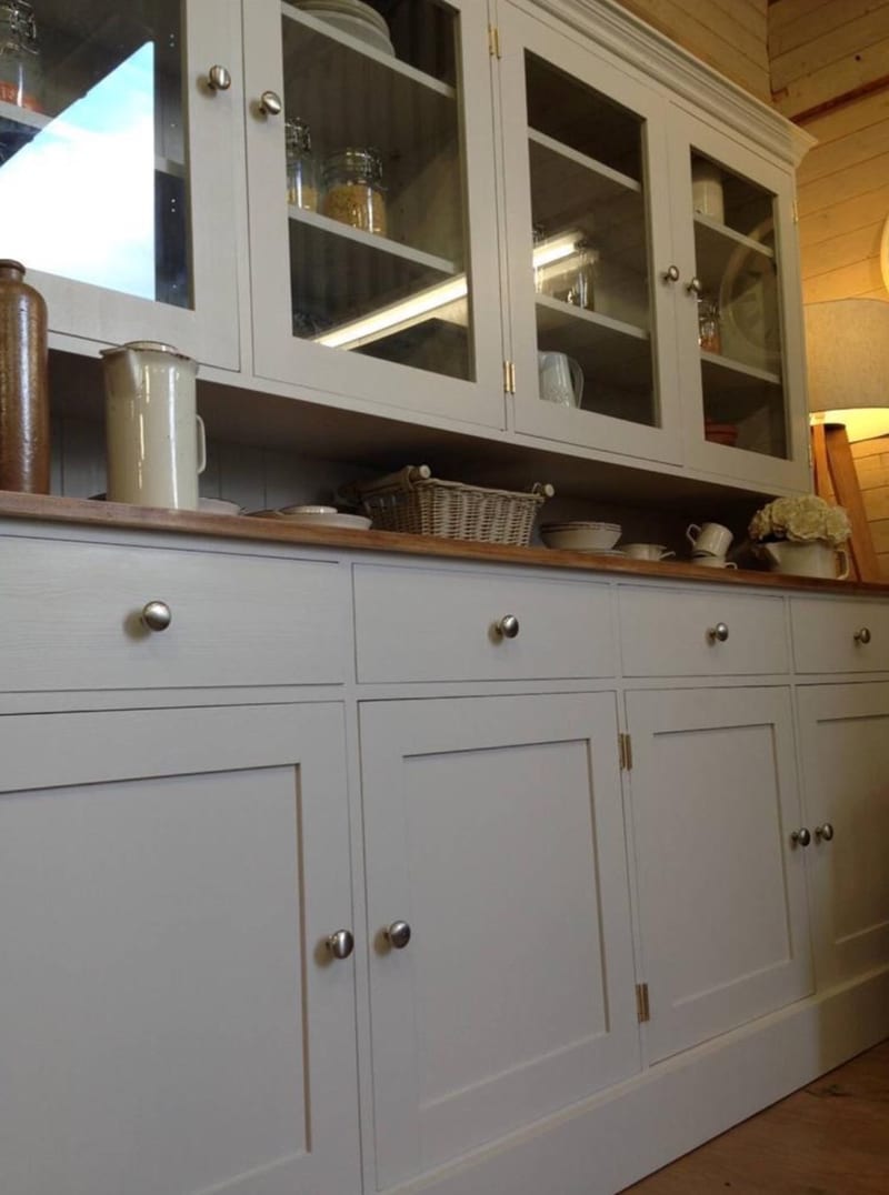 Angled view along the pale grey painted dresser showing four drawers and four cupboard doors with brushed nickel knobs, a waxed pine worktop and the glazed cabinet above