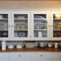 Front view of the glazed top of the dresser with four glass doors and brushed nickel knobs, shelves of storage jars and plates, and enamel pots and a wicker basket on the worktop below