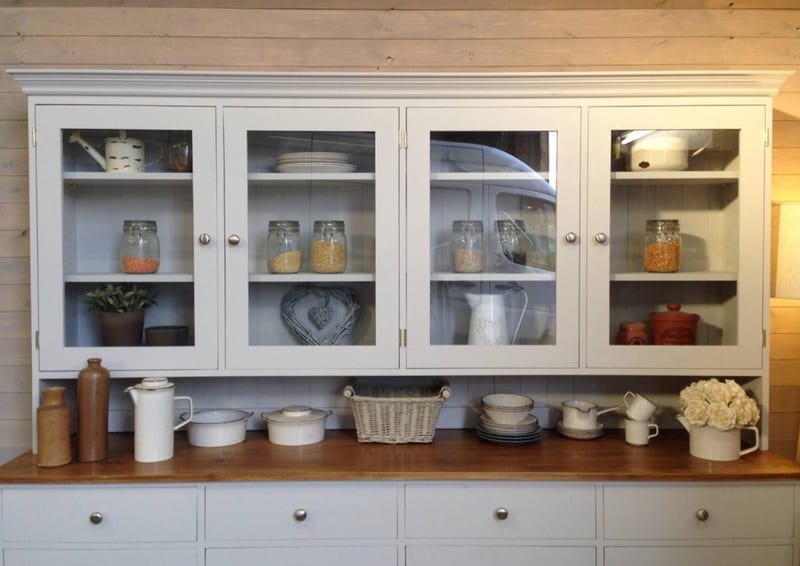 Front view of the glazed top of the dresser with four glass doors and brushed nickel knobs, shelves of storage jars and plates, and enamel pots and a wicker basket on the worktop below