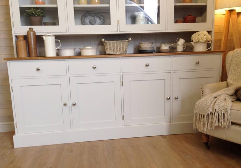 Angled view of the dresser base showing four drawers over four panelled doors with brushed nickel knobs, the waxed pine worktop and deep plinth, with a cream armchair on wood flooring