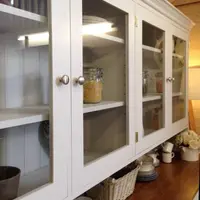 Angled close view of the glazed upper cabinet showing brushed nickel knobs, painted tongue and groove backboards, lit adjustable shelves and glass jars of pasta and pulses