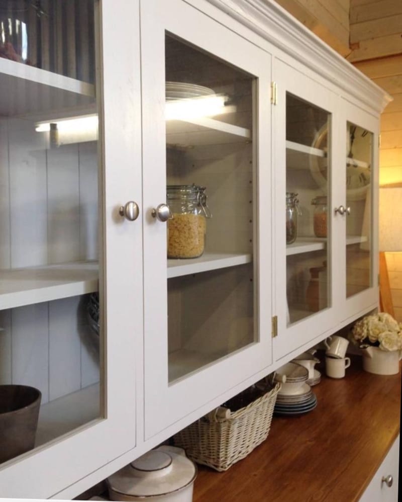 Angled close view of the glazed upper cabinet showing brushed nickel knobs, painted tongue and groove backboards, lit adjustable shelves and glass jars of pasta and pulses