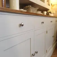 Low angled detail of the painted base showing the plain drawer fronts, a brushed nickel knob, brass butt hinges on the shaker doors and the rounded edge of the waxed pine worktop above
