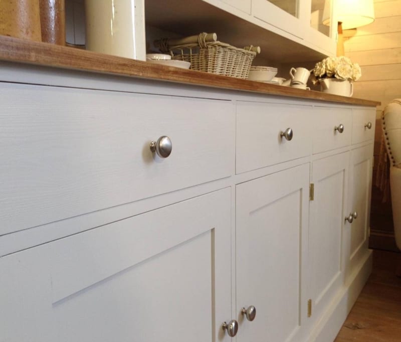 Low angled detail of the painted base showing the plain drawer fronts, a brushed nickel knob, brass butt hinges on the shaker doors and the rounded edge of the waxed pine worktop above
