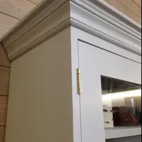 Close view of the top corner of the dresser where the moulded cornice meets the painted side panel, with a brass hinge on the glazed door and pine plank walls behind